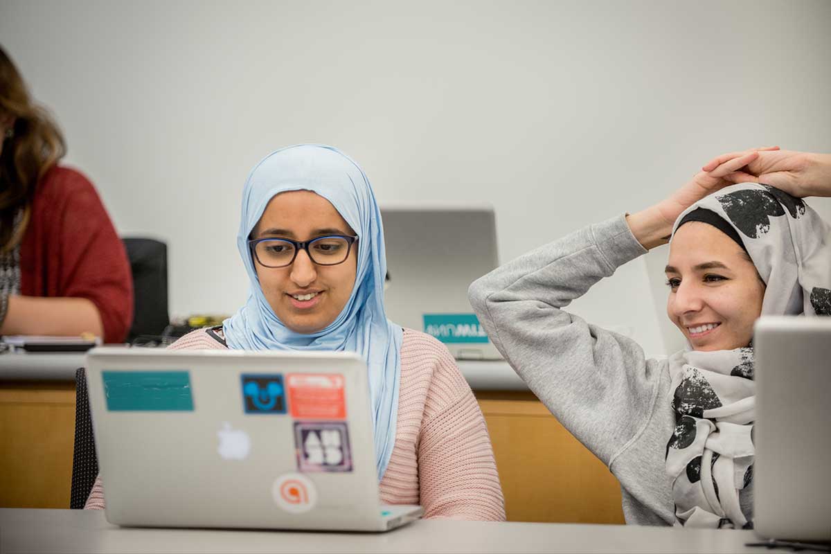 Two students view an open laptop while they sit in a classroom waiting for a class session to begin.