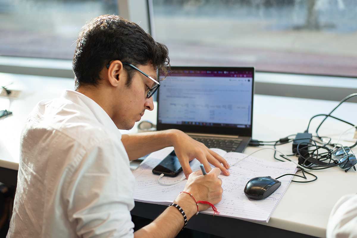 A student sitting at a table writes in a notebook. The student has a laptop open on the table in front of him. There is also a mouse and a tangle of cables on the table.