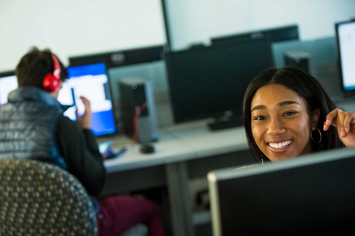 A student seated at a desk in a computer lab smiles as she looks over her computer monitor.