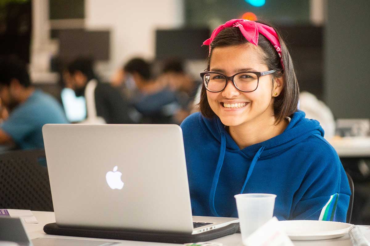 A student smiles as she sits at a table with her laptop open in front of her. Other students working on projects are visible in the background.