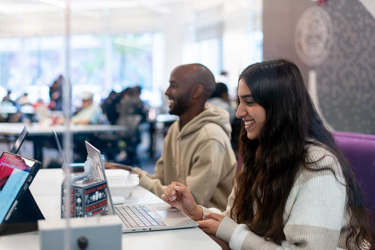 In West Village H's 102 lab, two students sit on a cushioned bench with their laptops open on the table in front of them. Both students are smiling.