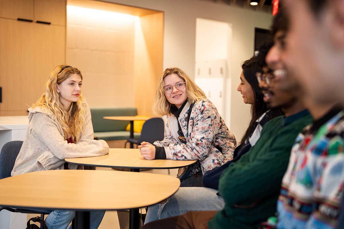 Six Northeastern students sit around two circular tables pushed together in a Northeastern meeting space.