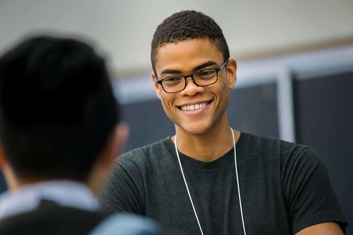 A Northeastern student smiles as he converses with another student. The back of the second student's head is visible in the foreground.