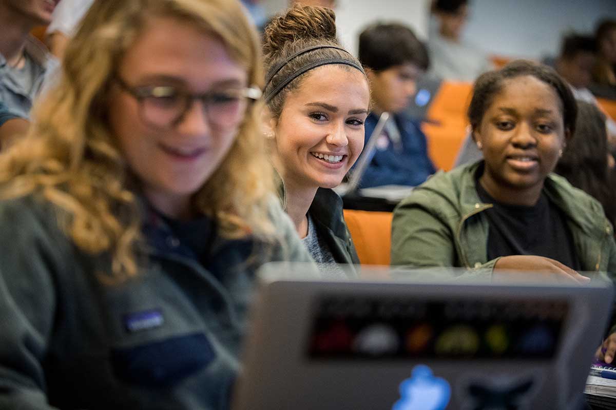 A group of students view a laptop screen during a class session in a Northeastern lecture hall. The room has orange seats. The three students in the foreground are grinning.