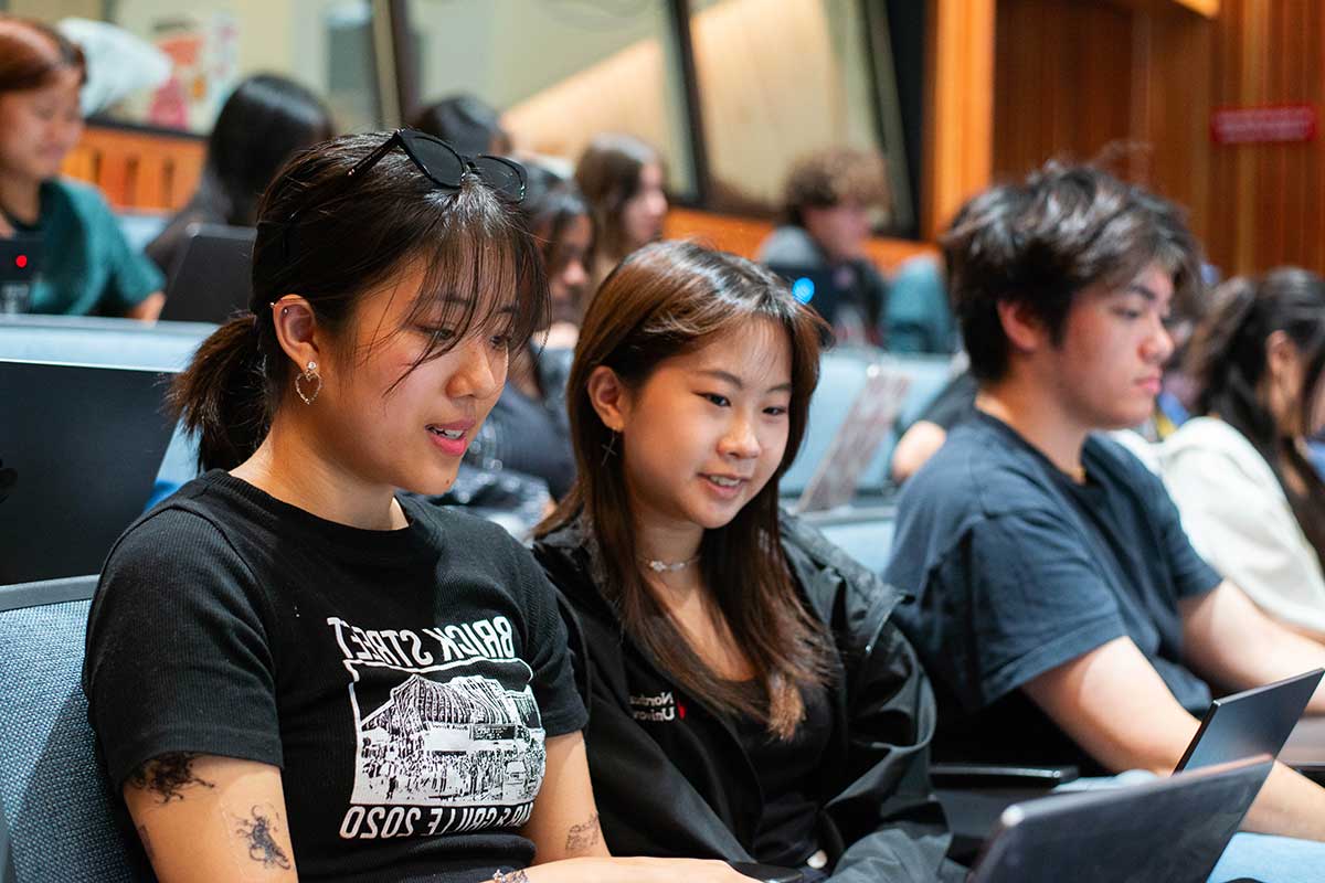 Several students sit in blue chairs during a class session in a Northeastern lecture hall. In the foreground, two students are viewing their laptops as they discuss their work.