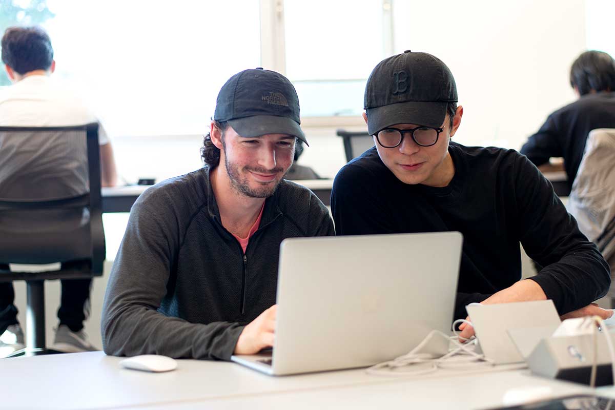 Two students sit at a table in the West Village H atrium viewing a laptop. The students are smiling while they work on a project. In the background, two more students are visible sitting in chairs as they work.