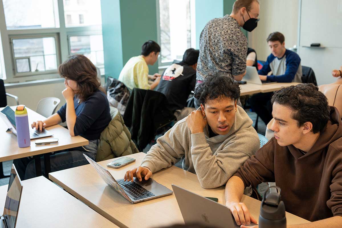 A Northeastern faculty member walks around a classroom as students sitting at tables work on a project. In the foreground, two students with laptops open in front of them are having a conversation.