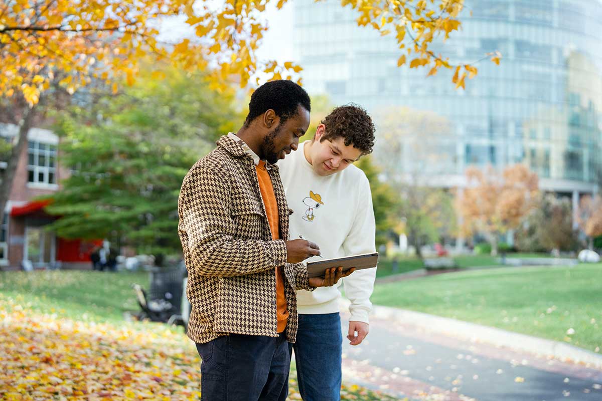 Two students stand in a Northeastern courtyard with trees with yellow leaves in the background. The student on the left is holding a tablet and stylus while the student on the right looks at the tablet.