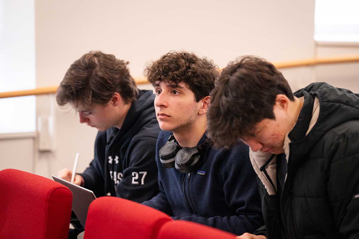 Three students sitting in a row of red chairs listen to a class lecture. The student on the right is writing with a pen, the student in the middle is looking forward, and the student on the right is viewing a mobile device.