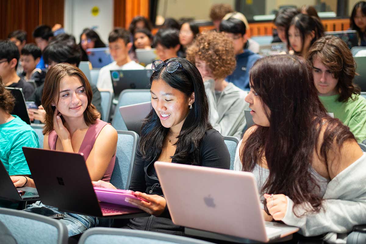 Dozens of students sit in blue chairs during a class session in a Northeastern lecture hall. In the first row, three students converse while they sit with their laptops open in front of them.