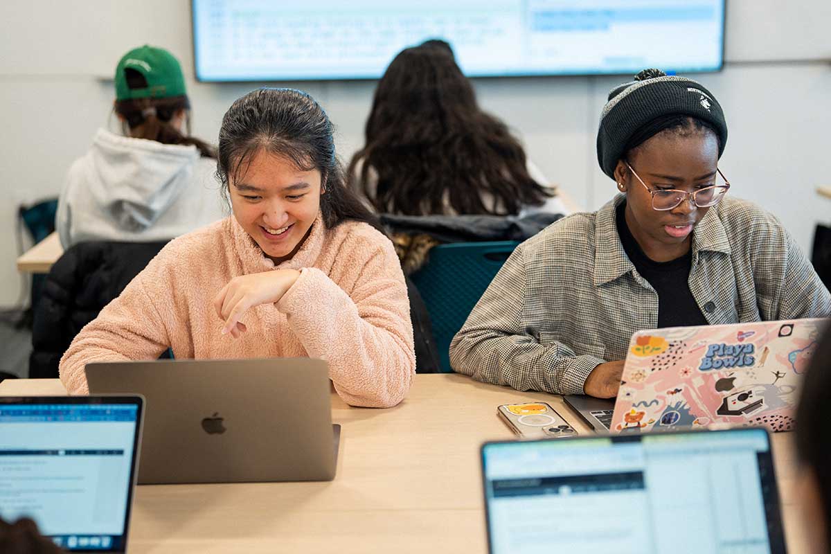 Two students sit at a table working on their laptops. The student on the left is smiling and the student on the right is typing.