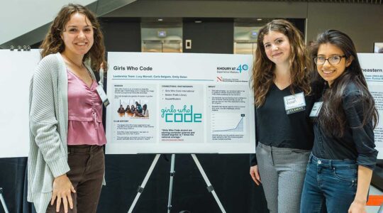 Members of Girls Who Code Boston stand around their club poster in Northeastern's ISEC Building. One member stands to the left of the poster. Two other members stand on the right side. The poster has sections for the club's mission, history, partnerships, and impact.