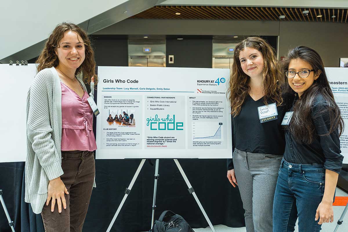 Members of Girls Who Code Boston stand around their club poster in Northeastern's ISEC Building. One member stands to the left of the poster. Two other members stand on the right side. The poster has sections for the club's mission, history, partnerships, and impact.