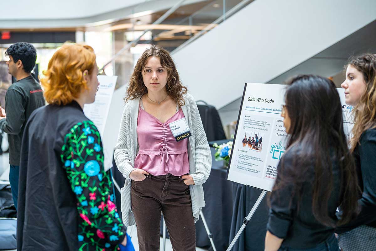 Three members of Girls Who Code Boston stand around their club's poster at a club fair in Northeastern's ISEC building. One person asks the club members a question.