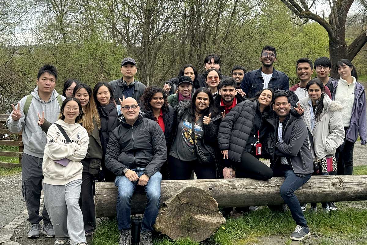 22 members of Khoury College Social Club. pose for a photo at a trailhead. Three of the members are sitting on a large log while the other members stand behind the log.