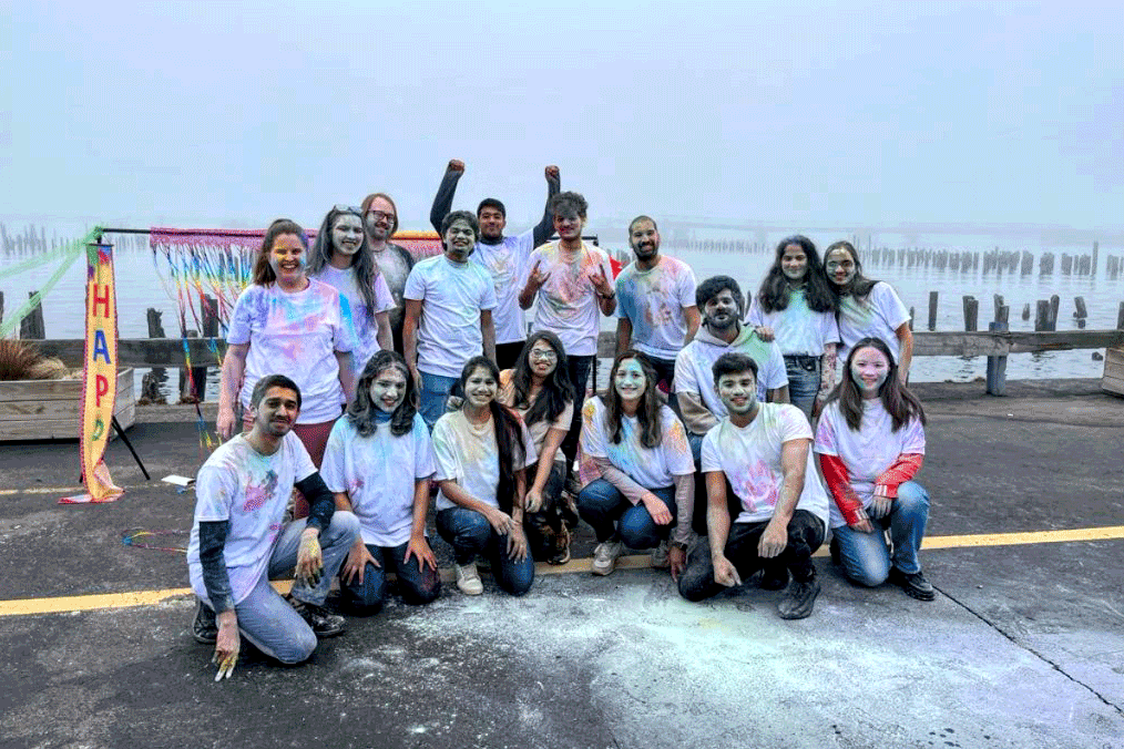 17 members of Namaste pose for a photo while covered in colorful powder after a color run. Eight members are kneeling in the front row. Nine students are standing in the back row.