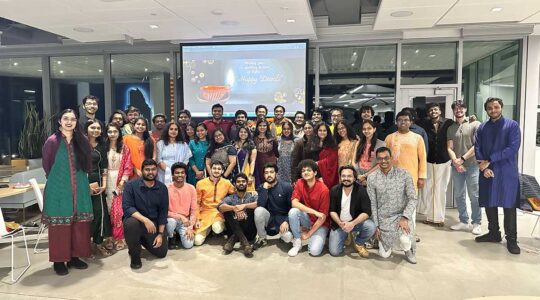 41 members of Namaster pose for a photo at the club's Diwali celebration. In the background, a projection screen shows a message that says, "Happy Diwali".