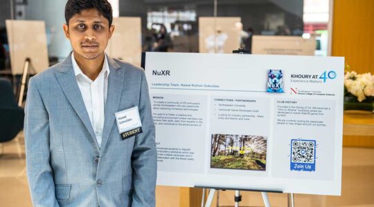 A member of NuXR stands to the left of his club's poster during a club fair. The poster shows the club's mission, partnerships, and history.