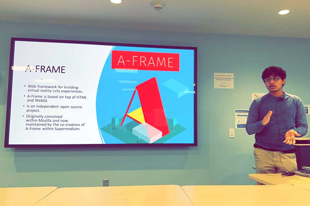 A member of NuXR speaks during a PowerPoint presentation. A large screen shows the presentation on the left side of the photo. The screen shows information about the A-Fram web framework. At the right of the photo, the student is speaking.