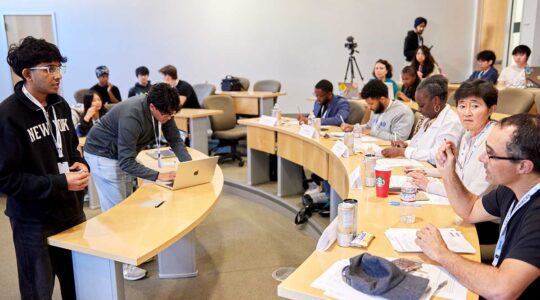 Members of Pawhacks present a project in a classroom. Two members of the club are standing at the front of the classroom. There are five other students and faculty members sitting at a row of desks. The faculty member in the foreground is asking a question while holding a pen.