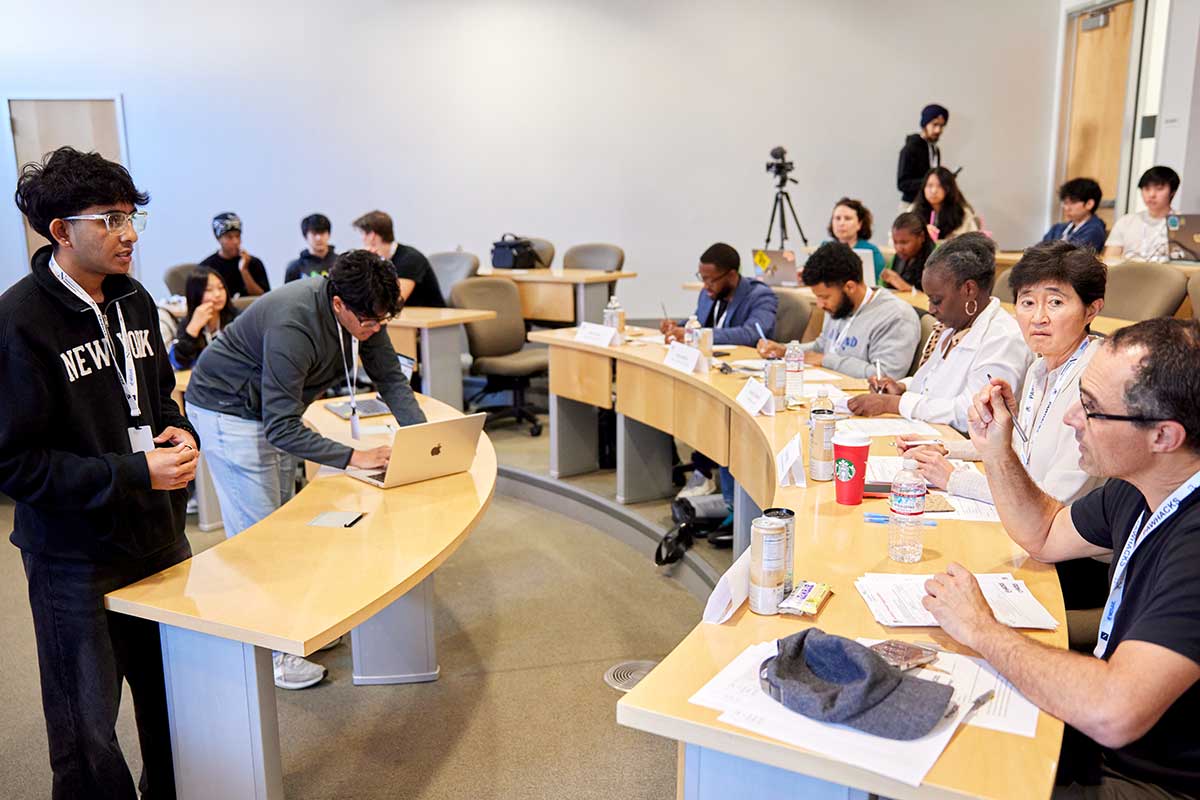 Members of Pawhacks present a project in a classroom. Two members of the club are standing at the front of the classroom. There are five other students and faculty members sitting at a row of desks. The faculty member in the foreground is asking a question while holding a pen.