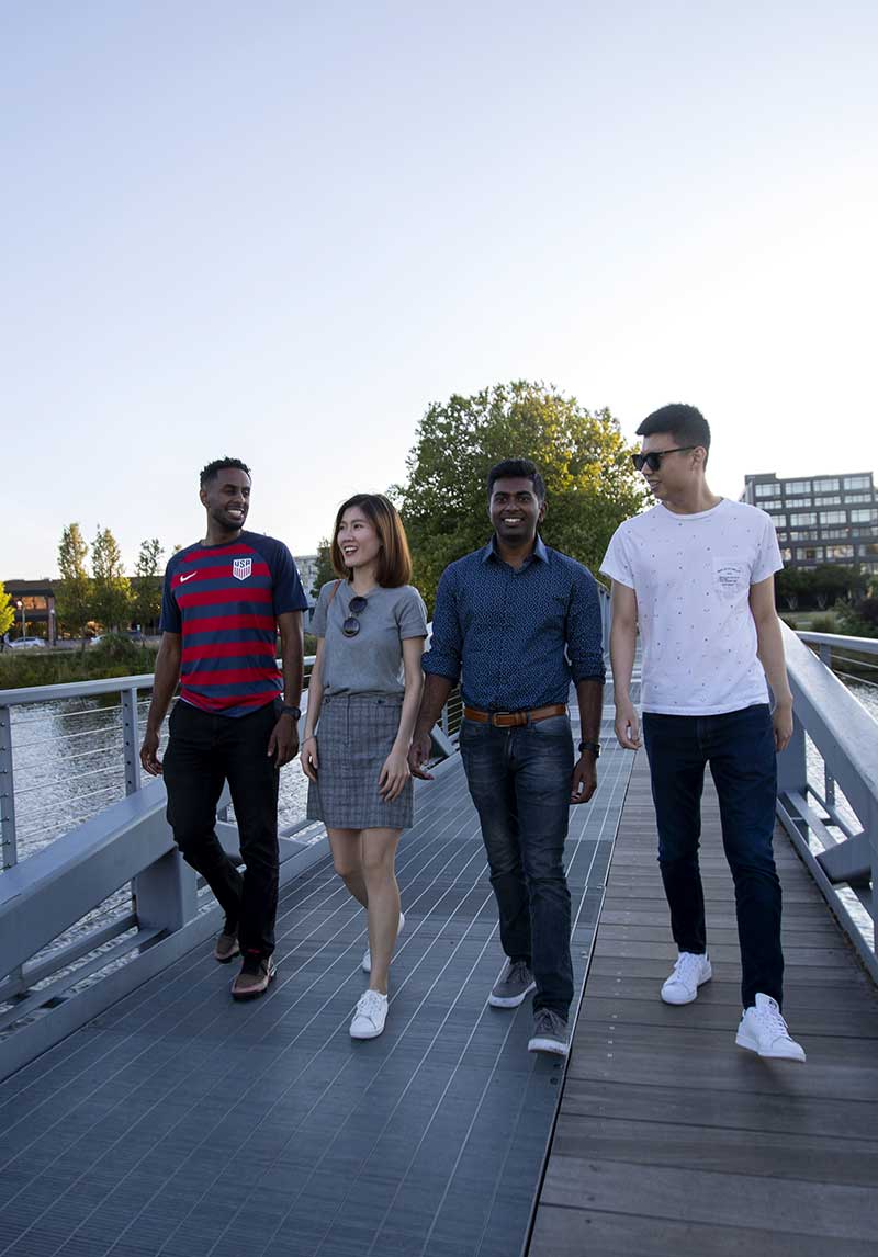 alt="Four students walk across a pedestrian bridge at Northeastern Seattle on a clear, sunny day. The Space Needle is in the background on the left side of the photo.