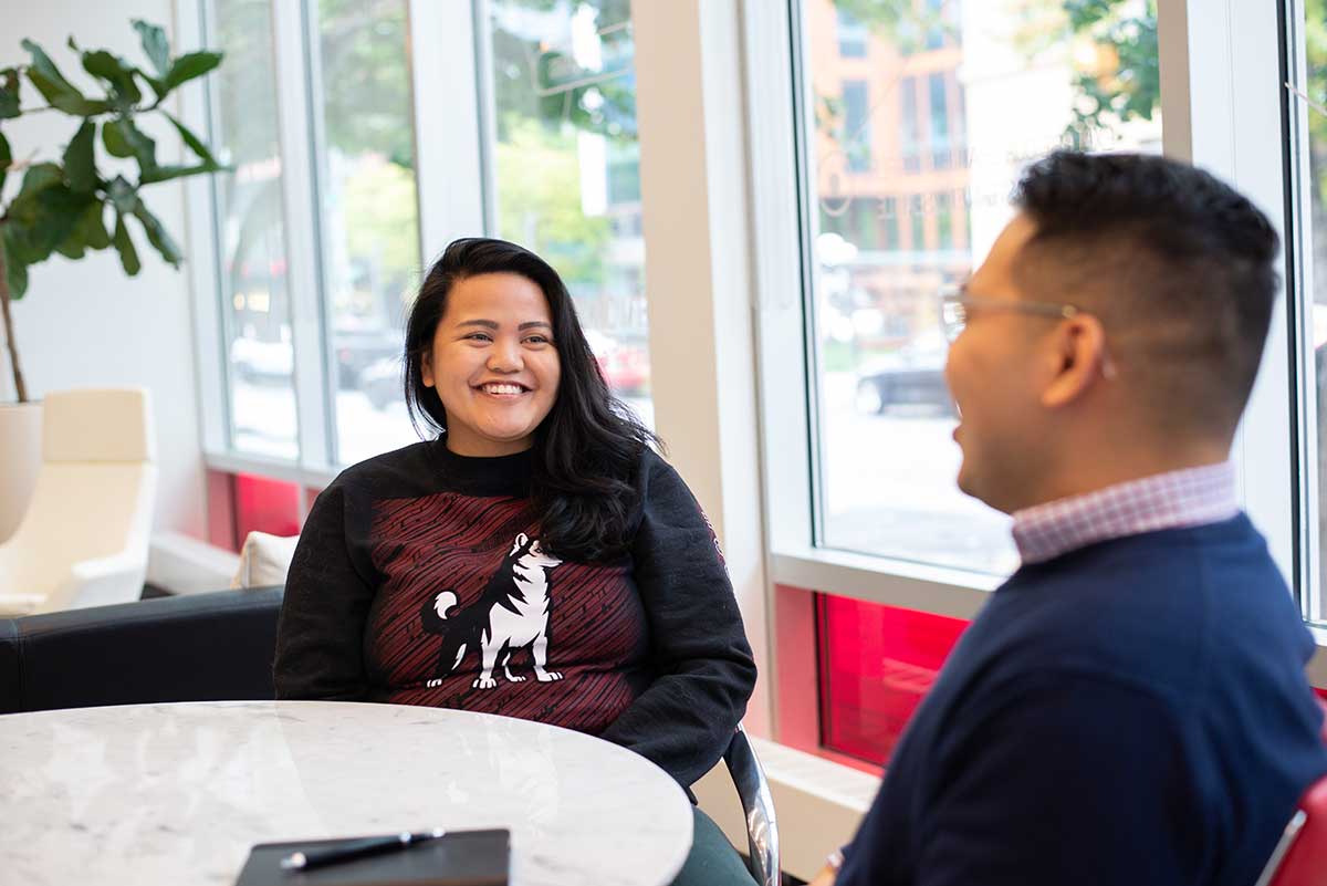 Two students laugh during a conversation at Northeastern Seattle. The students are sitting around a round, white table.