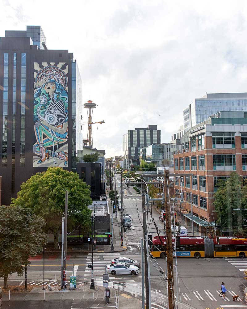 A photo shot from the top of Northeastern Seattle's building. In the distance, the side of a building is covered in a mural and the Space Needle can be seen farther in the distance. The sky is covered in white clouds.