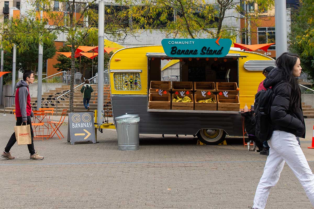 An exterior photo of Northeastern Seattle's campus. The photo shows a yellow food truck with a sign that says "Community Banana Stand." The stand has several wooden boxes filled with bananas and other red fruits. There are buildings of the left and right edges of the photo. Three people can be seen walking through the courtyard.