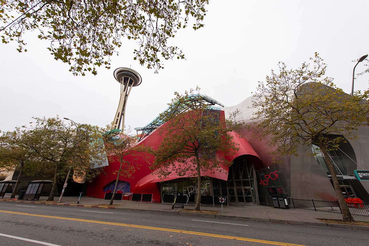 A photo of a building with a red and silver curved roof in Seattle. In the background, the Space Needle rises into the sky, which has gray clouds.