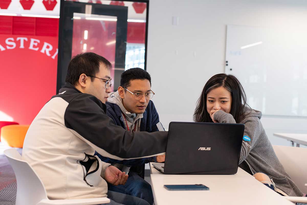 Three students sitting at a table look at one laptop. The student on the left side of the group is pointing at the laptop.