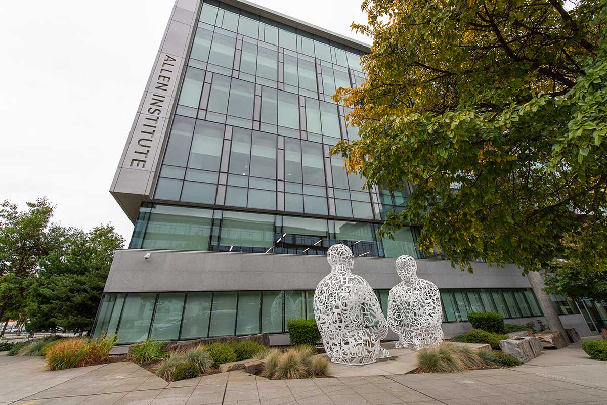 A photo of the Allen Institute building in Seattle. The buildings exterior is glass and stone, and a large tree with green leaves appears in the right side of the photo.