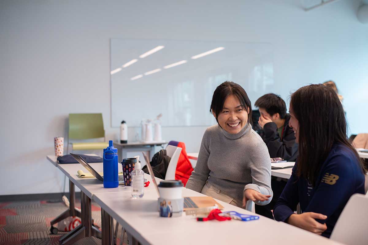 Two students talk during a class break in a classroom at Northeastern Seattle. The students are seated behind a table with their laptops open.