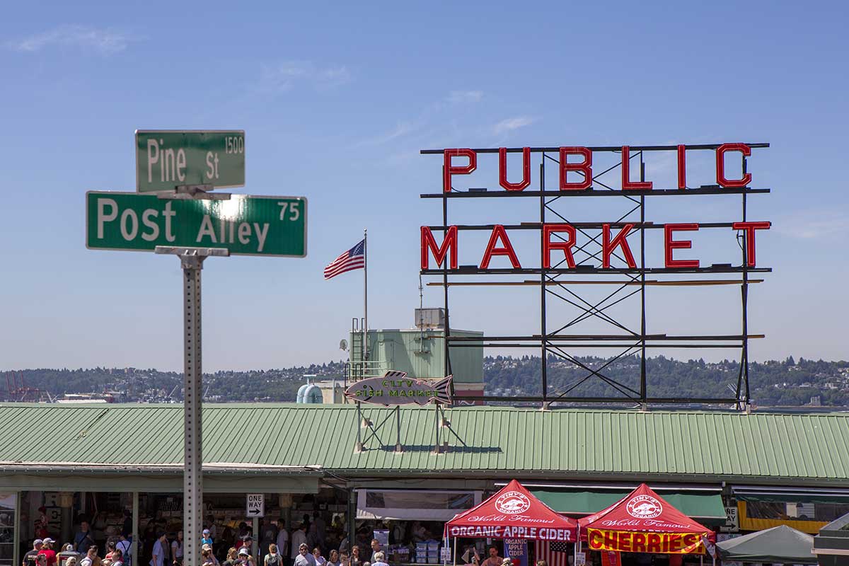 A photo of the large, red Public Market sign on the green roof of Pike's Place Market in downtown Seattle. The sign has all capital letters. A street sign marking the intersection of Pine St. and Post Alley are in the foreground. A blue sky with some white cloulds is visible in the background.