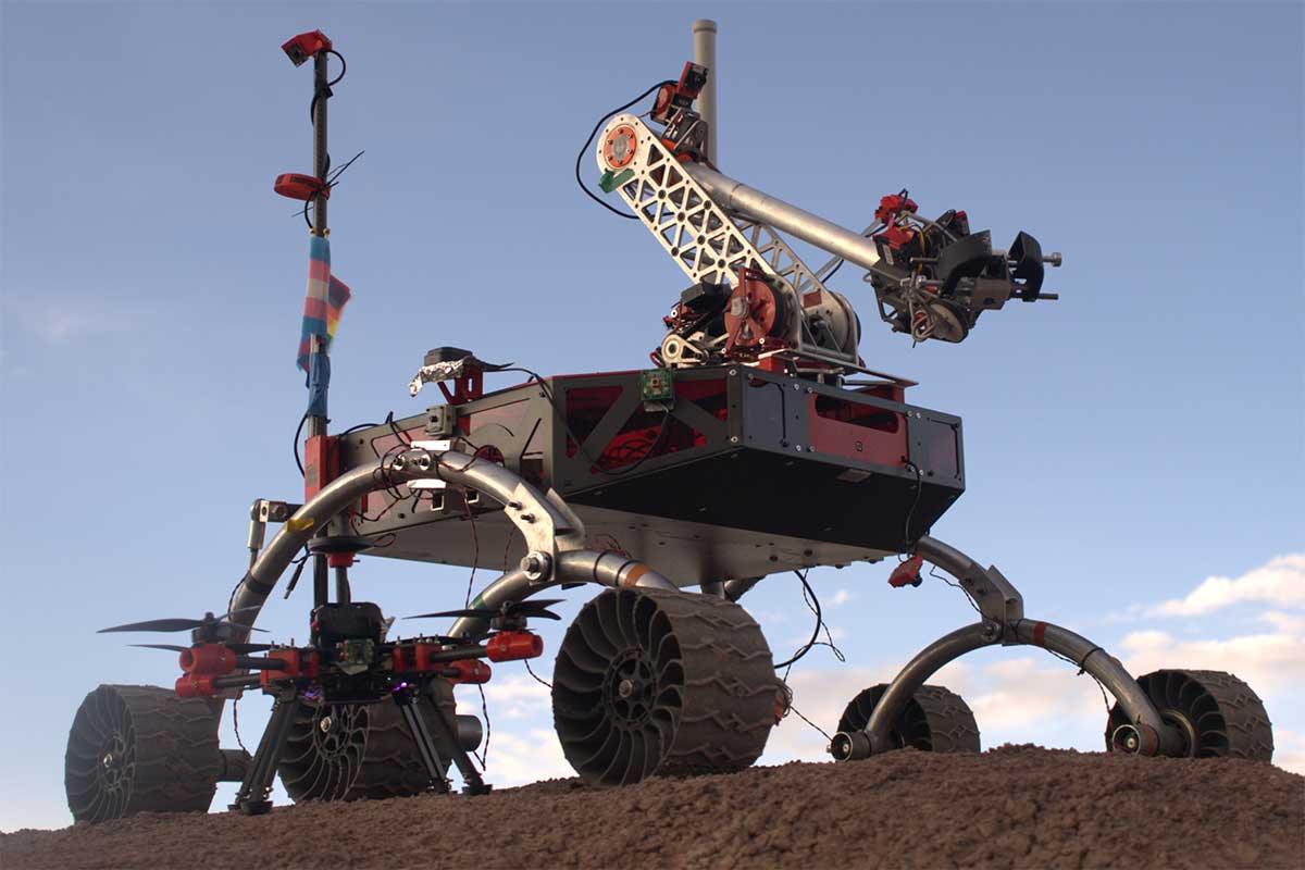 A rendering of a space vehicle on red, rocky terrain. The vehicle has three wheels on each side, for a total of six wheels. The front of the vehicle has a robotic arm with cameras at the end of the arm. An antenna is sticking up at the rear of the vehicle.