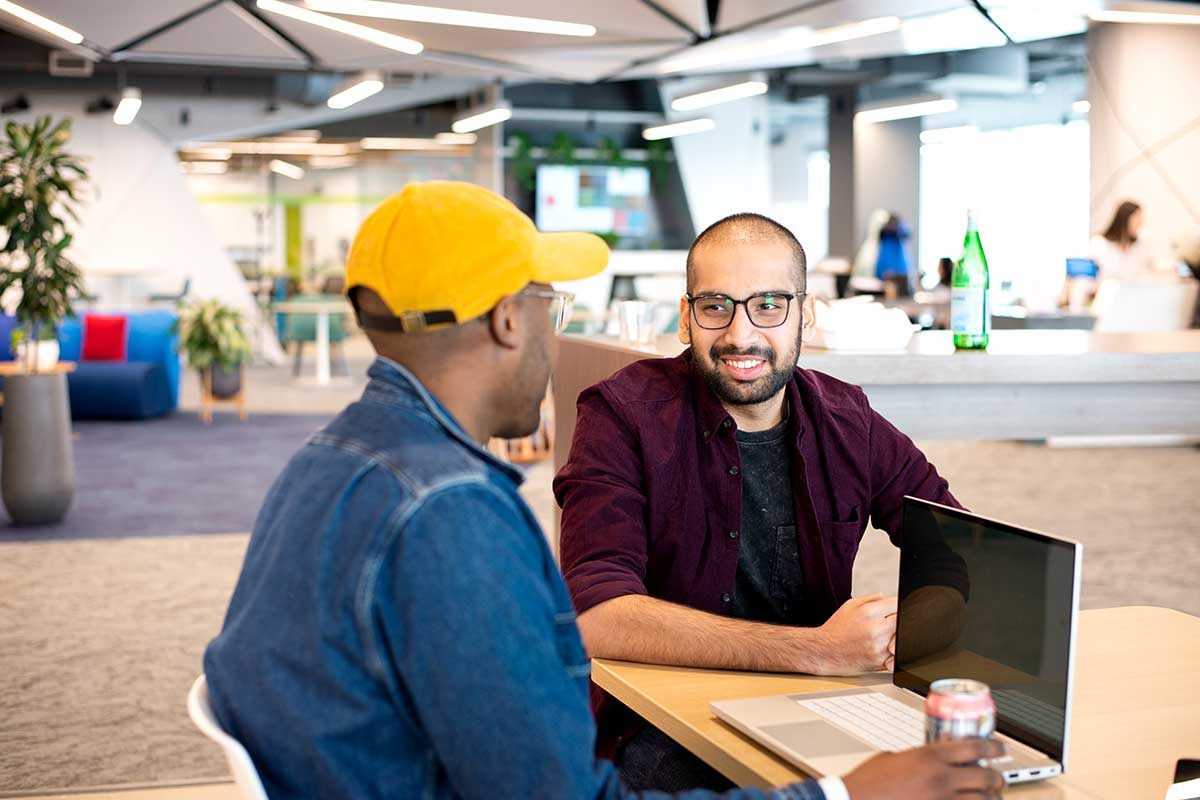 Two students discuss a project in a shared workspace at the Roux campus in Portland. The students are sitting in chairs at a rectangular table. One student has a laptop open.