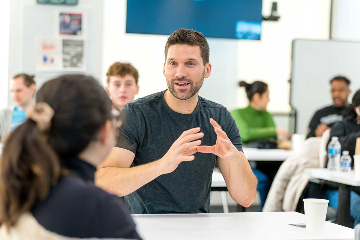 A Khoury student sitting on one side of a rectangular table talks with another student sitting across the table. The student who is speaking is gesturing with his hands by making a round shape with both hands. Other students sit around several other tables in the background.