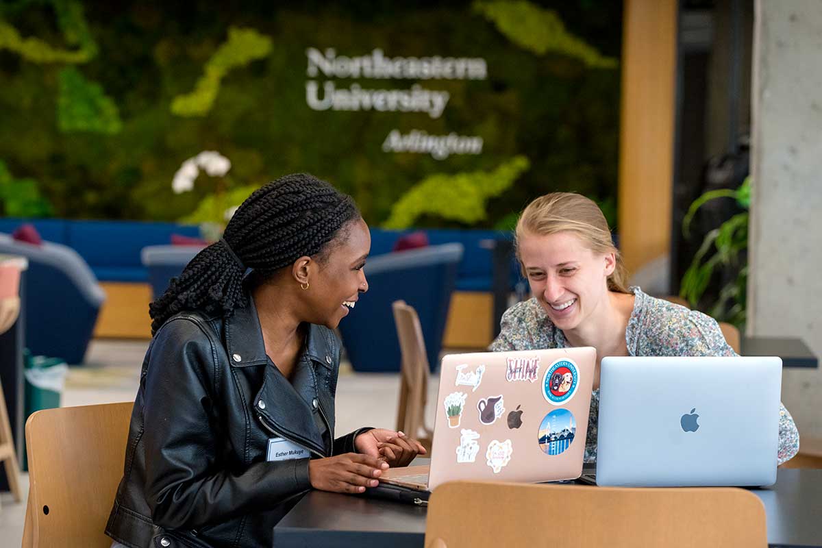 Two Northeastern Arlington students laugh as the view laptop screens in a campus lounge.