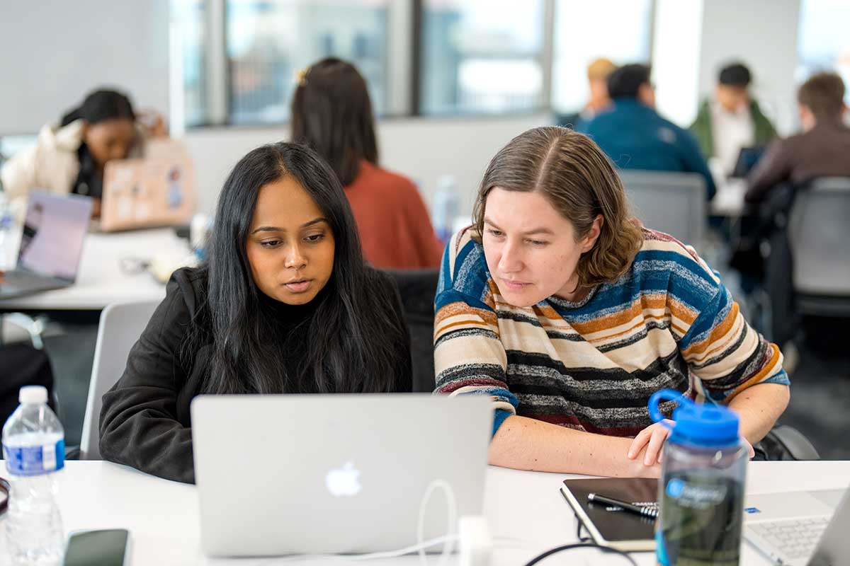 Two students sit at a table viewing a shared laptop screen. In the background, other groups of students are sitting around circular tables.