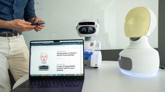 An open laptop sits on a desk alongside a robot that resembles a human head and neck