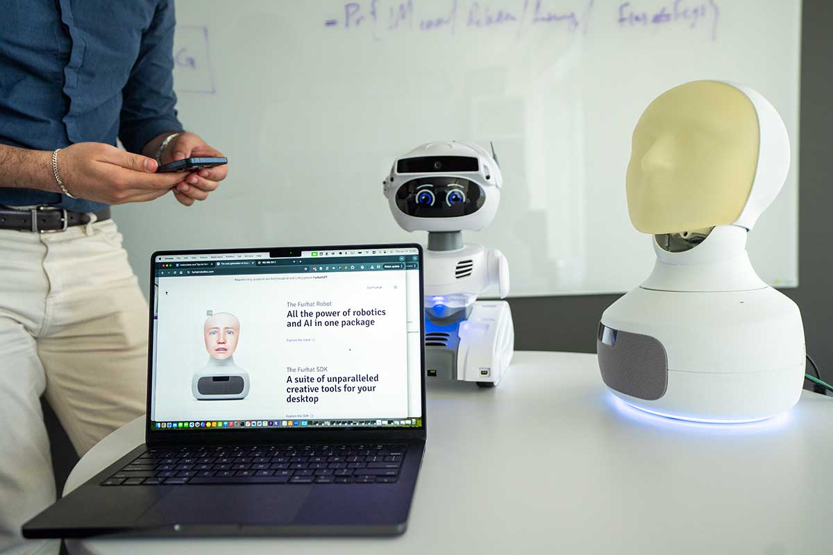 An open laptop sits on a desk alongside a robot that resembles a human head and neck