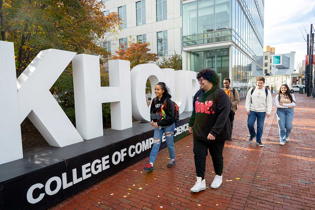 Students walk by West Village H on the Boston campus. A large, white KHOURY sign is next to the students. West Village H rises in the background.