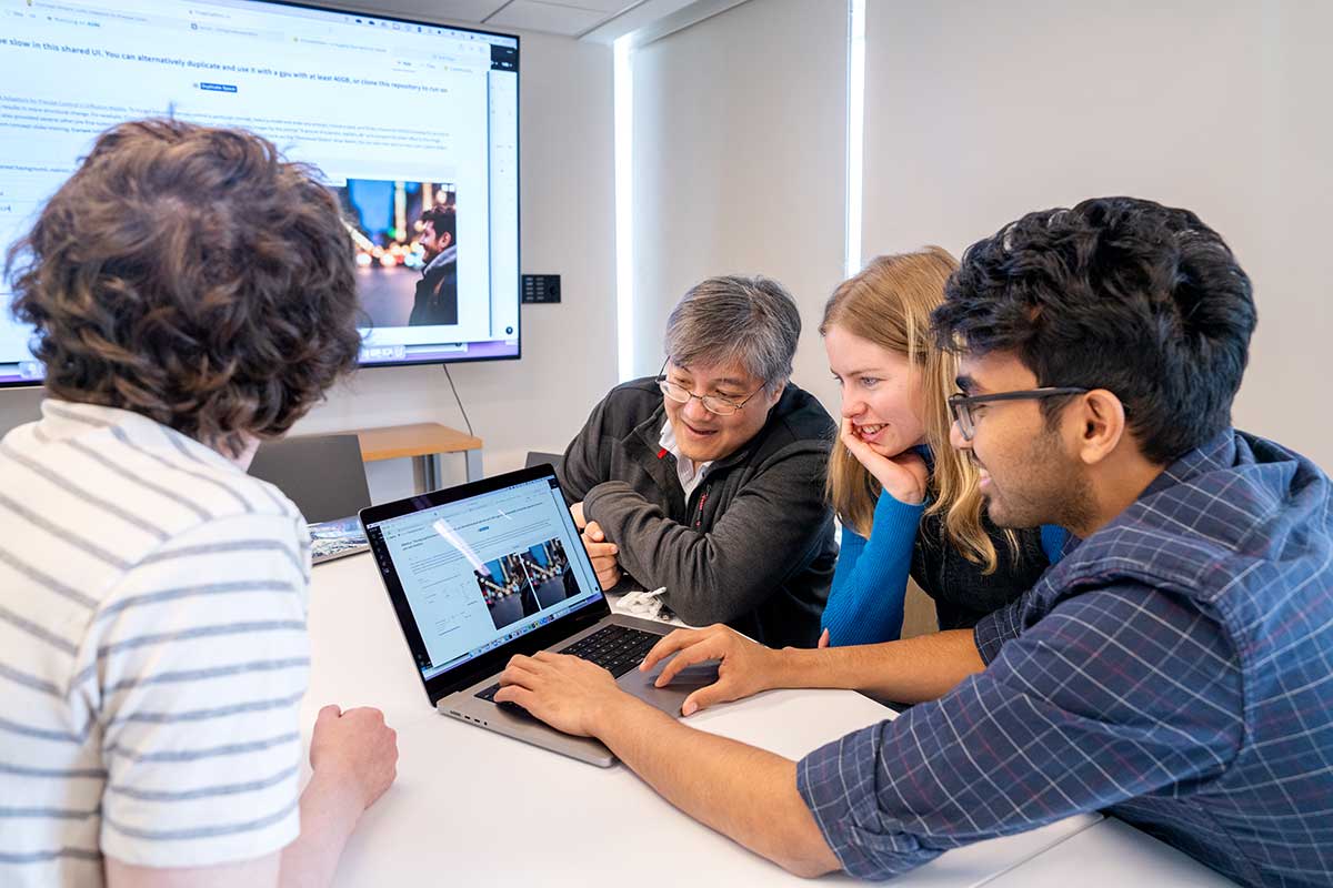 Faculty member DAvid Bau, second from left, views a laptop screen with a group of students. Two students are sitting on Bau's right, and another is setting on Bau's left.