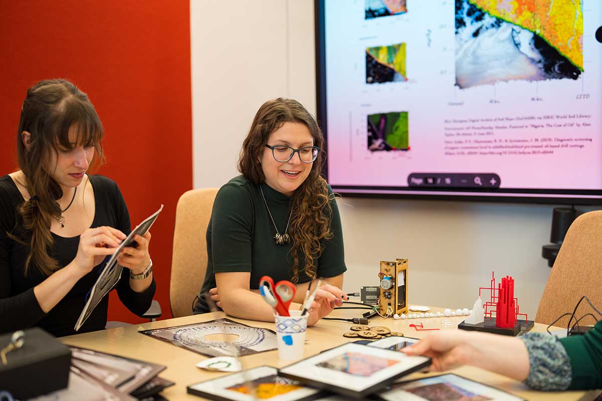 Two students work collaboratively while sitting at a table in a Khoury conference room. There is a large screen with several data visualizations on the room's wall.