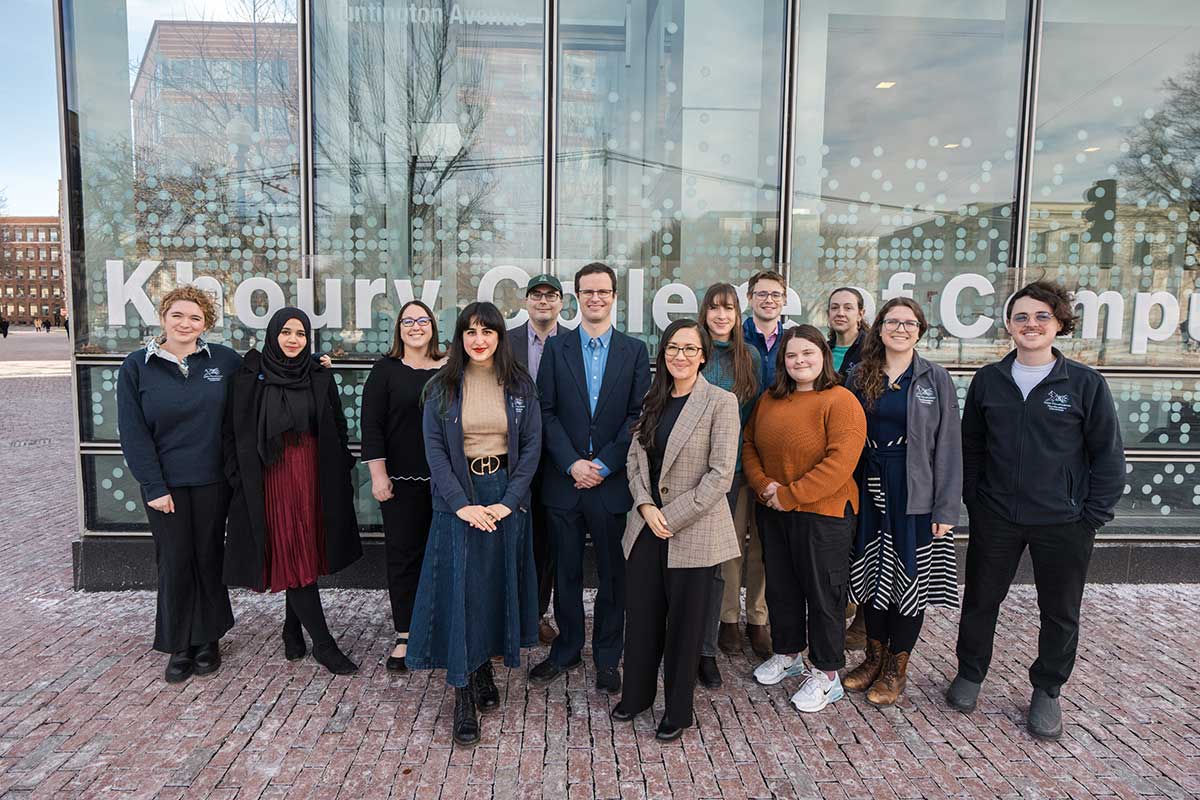 A group of thirteen data visualization researchers at Khoury College stand outside the West Village H building and pose for a photo. The building's glass exterior is in the background.