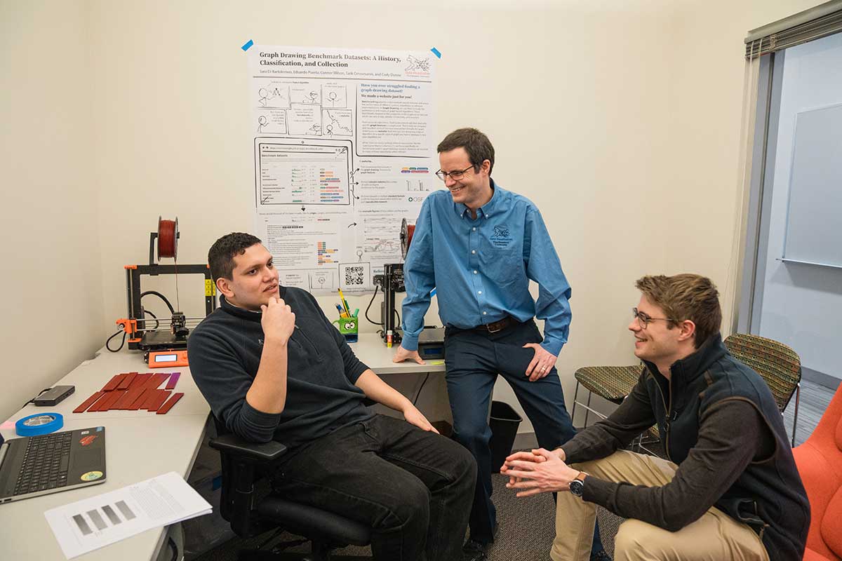 A Khoury faculty members sits on a desk and smiles while one student to his left, who is sitting in a chair, talks about his work. Another student sitting in a chair to the right of the faculty member smiles.