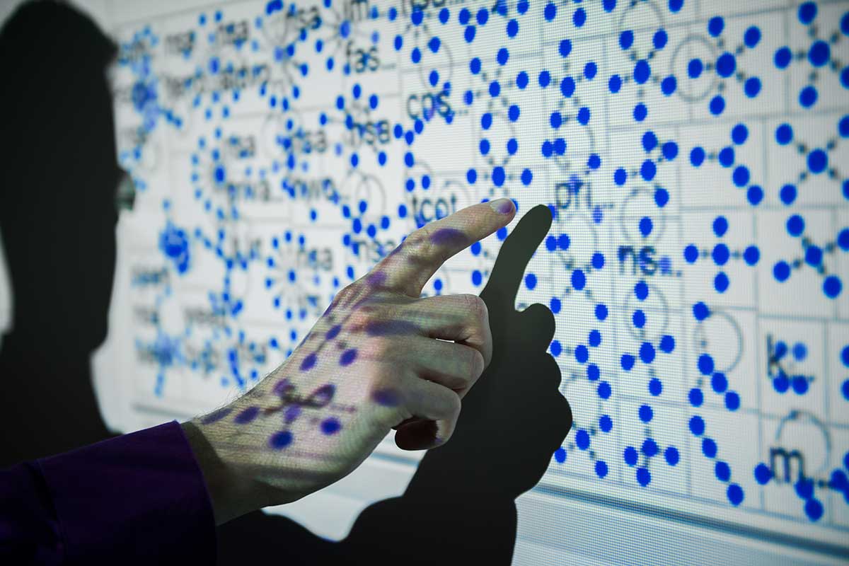 An unseen face points at a data visualization composed of many blue dots that is projected on the wall. We see the person's hand pointing, as well as the shadow of the and and person's head on the screen.