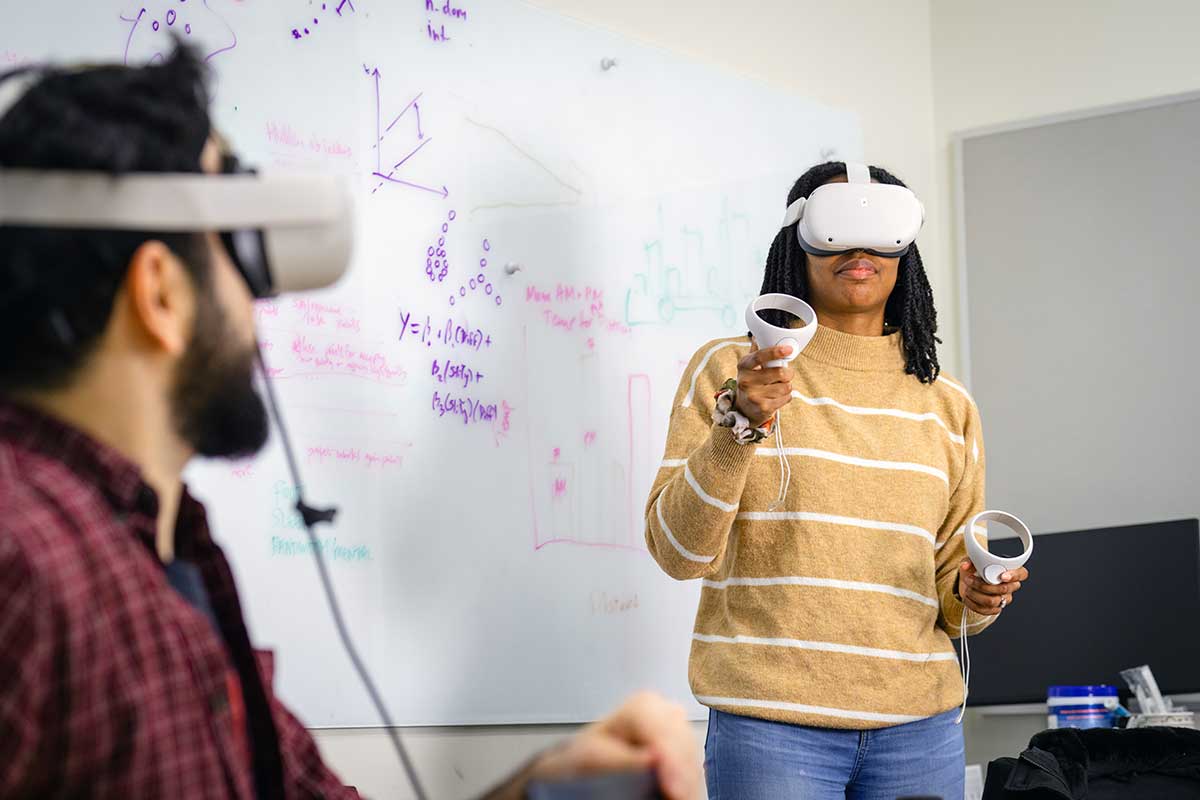 Two student wear virtual reality headgear while playing a game. One student is also holding two controllers shaped like cones.