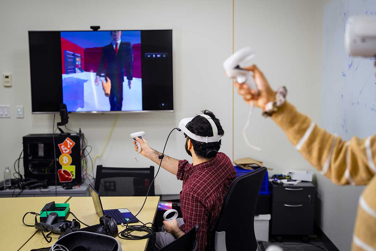 Two students wearing virtual reality goggles hold a game controller as a character moves on a television screen. The television is attached to the room's white wall.