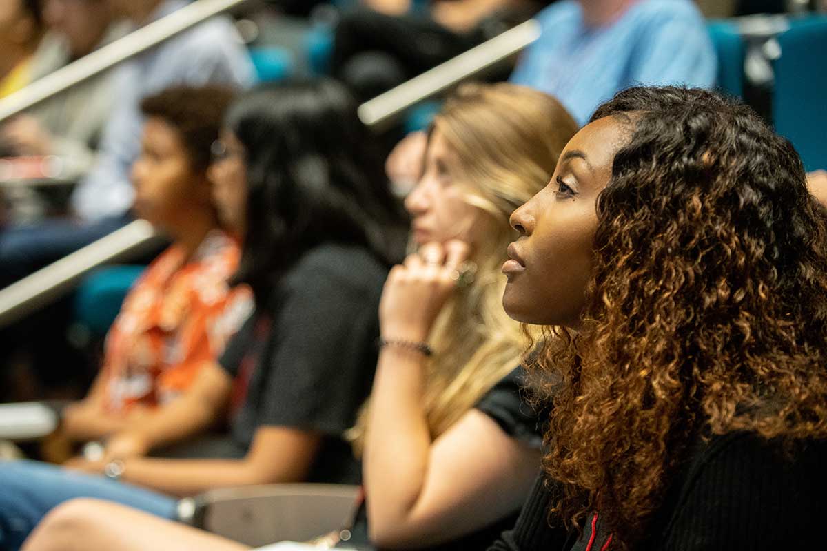 Four students sit in a row of seats in a lecture hall. The student closest to the camera is looking intently at the front of the hall.